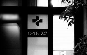 Sign with medical cross symbol reading 'Open 24h' on a glass door