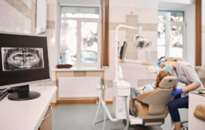 Dentist wearing mask and gloves examines a patient in a dental chair, with a dental X-ray displayed on a nearby monitor