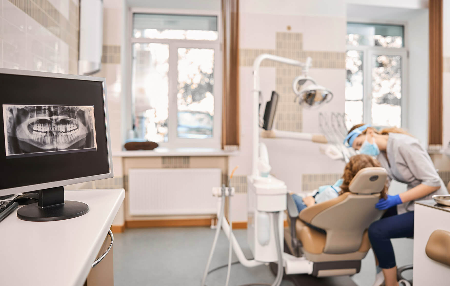 Dentist wearing mask and gloves examines a patient in a dental chair, with a dental X-ray displayed on a nearby monitor