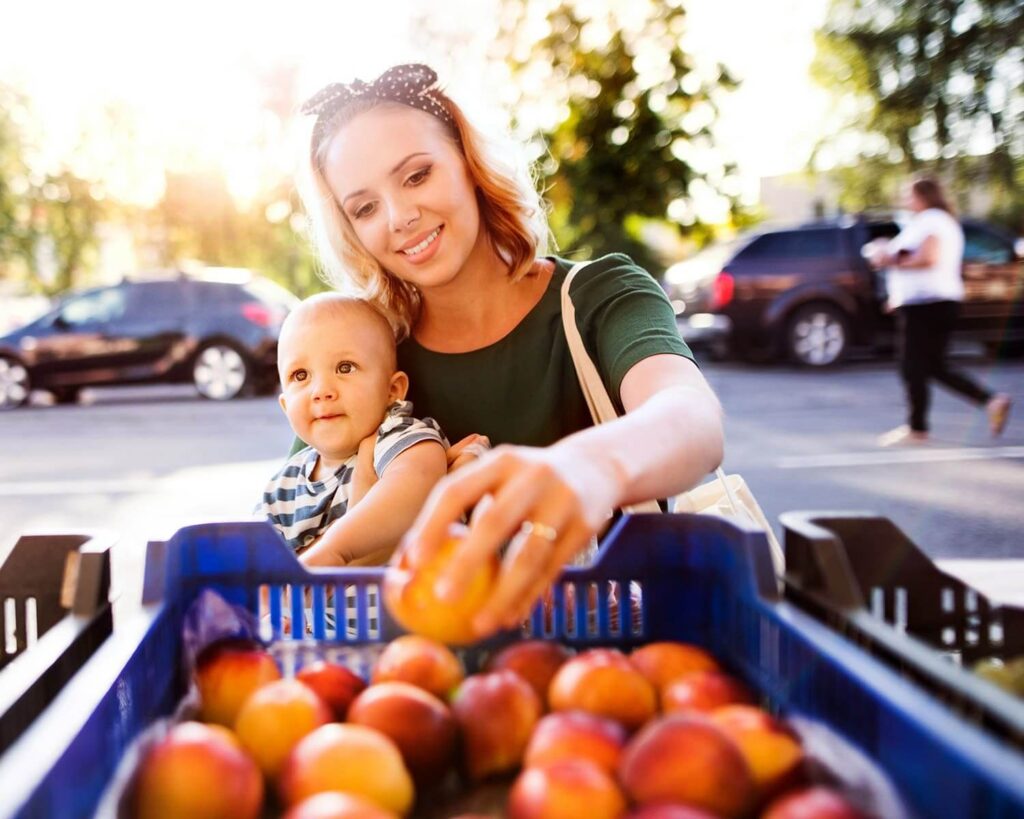 Smiling mother holding baby while picking fresh peaches at a farmers market.
