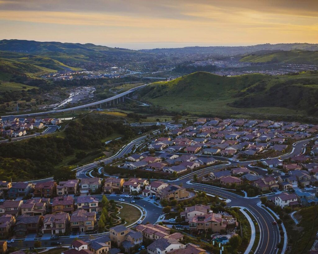 Large location pin icon over aerial view of Mission Viejo CA neighborhoods and winding roads surrounded by rolling green hills at sunset