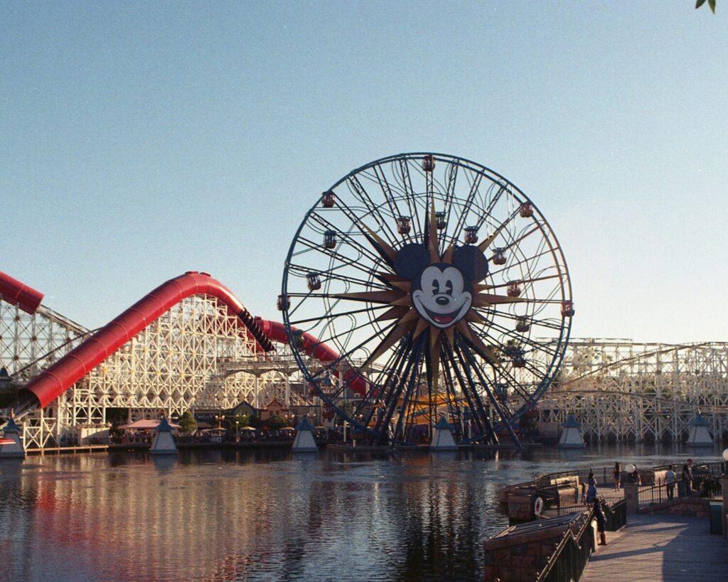 Ferris wheel with Mickey Mouse face at Disney California Adventure Park beside a lake, with roller coaster and boardwalk in Anaheim, California