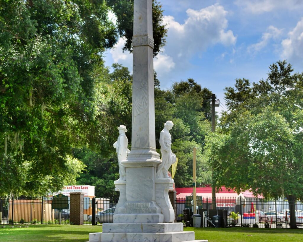 Marble obelisk monument with statues at its base in a sunny park, green trees and a fence behind. A market or fair is visible in the background.
