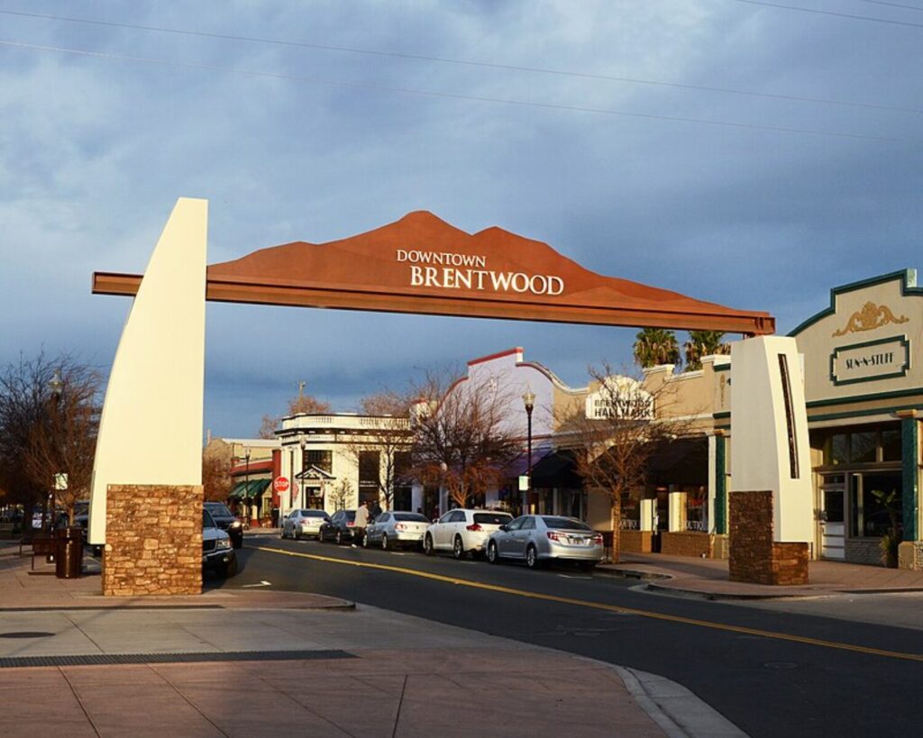 Street view of downtown Brentwood, California with an archway sign over the road, lined shops, and parked cars under a cloudy sky