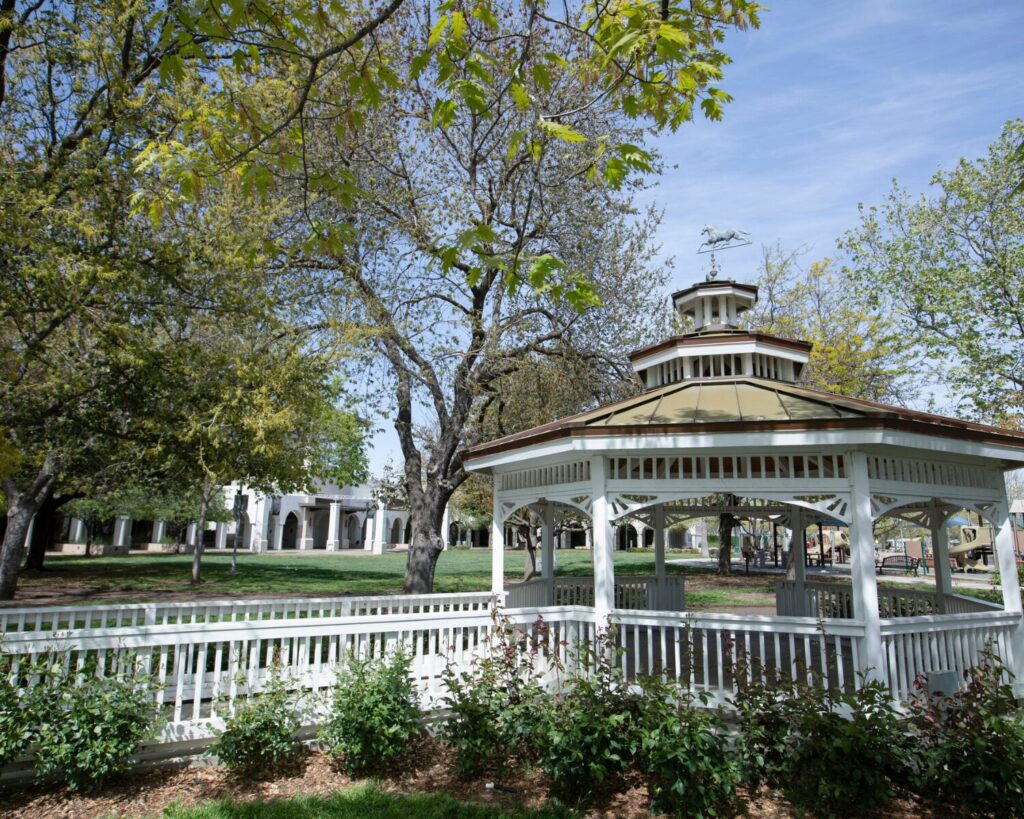 White gazebo in a green park in downtown Brentwood, California, surrounded by trees and a wooden fence under a clear blue sky