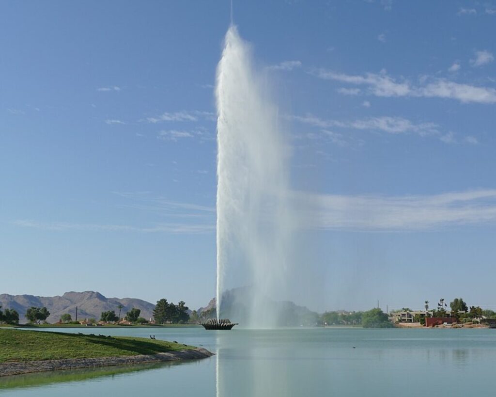 Large water fountain shooting a tall vertical jet at full capacity in the middle of a lake, with calm water, grassy shoreline, distant mountains, and a clear blue sky in the background.