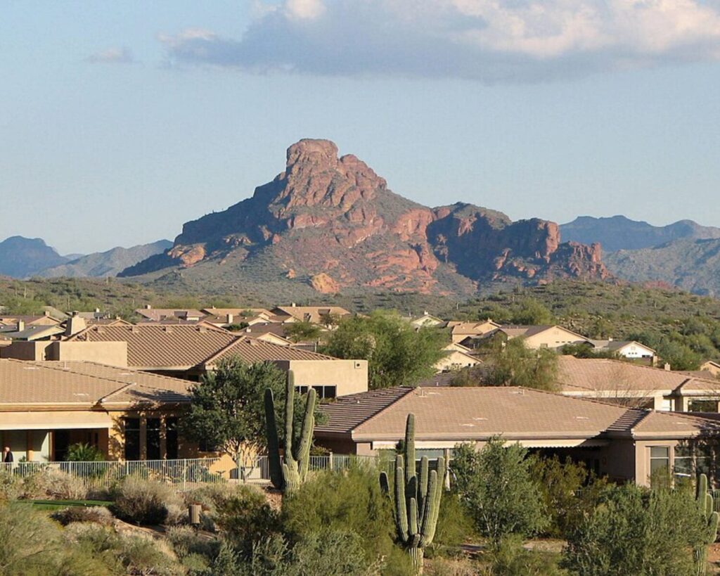 Residential neighborhood in Fountain Hills, Arizona, with desert-style homes and cacti in the foreground and a rugged red rock mountain rising in the background under a partly cloudy sky.