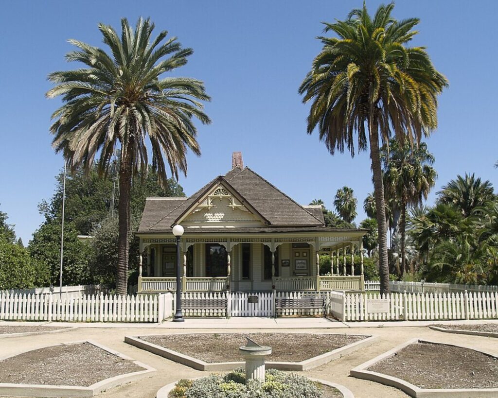 Historic Dr. George Clark House with a front porch, surrounded by tall palm trees and a white picket fence under a clear blue sky