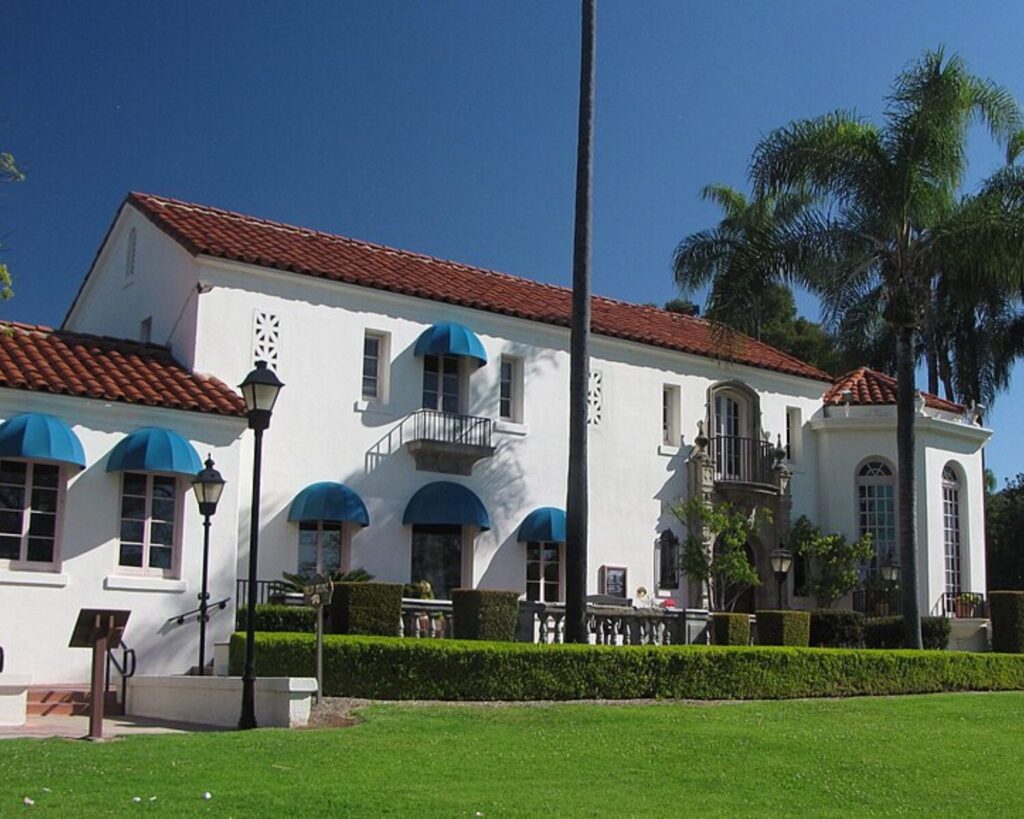 Muckenthaler House, a white Spanish-style mansion with red tile roof, blue awnings, and palm trees in a landscaped garden