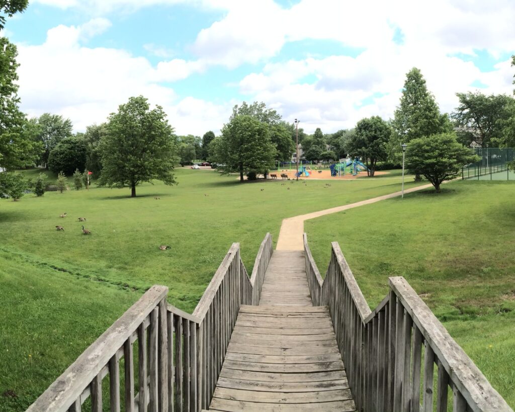 Panoramic view of Schaumburg Illinois park with wooden bridge pathway green lawns trees and open recreational landscape USA