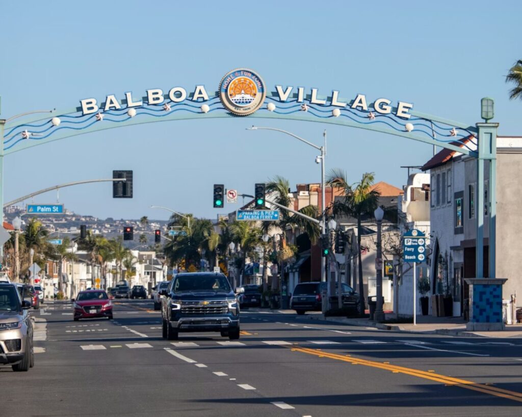 Street view of Balboa Village entrance arch in Newport Beach with cars driving along a palm-lined road