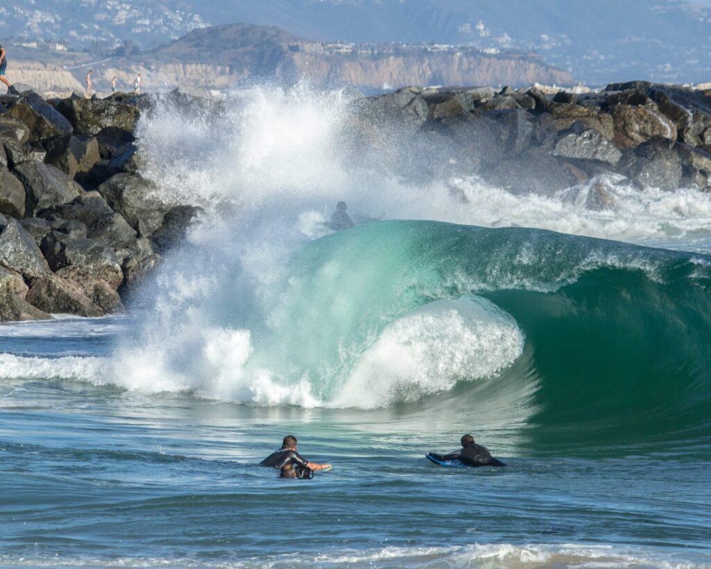 Surfers paddling in the ocean as a powerful curling wave crashes near rocky shoreline during daytime
