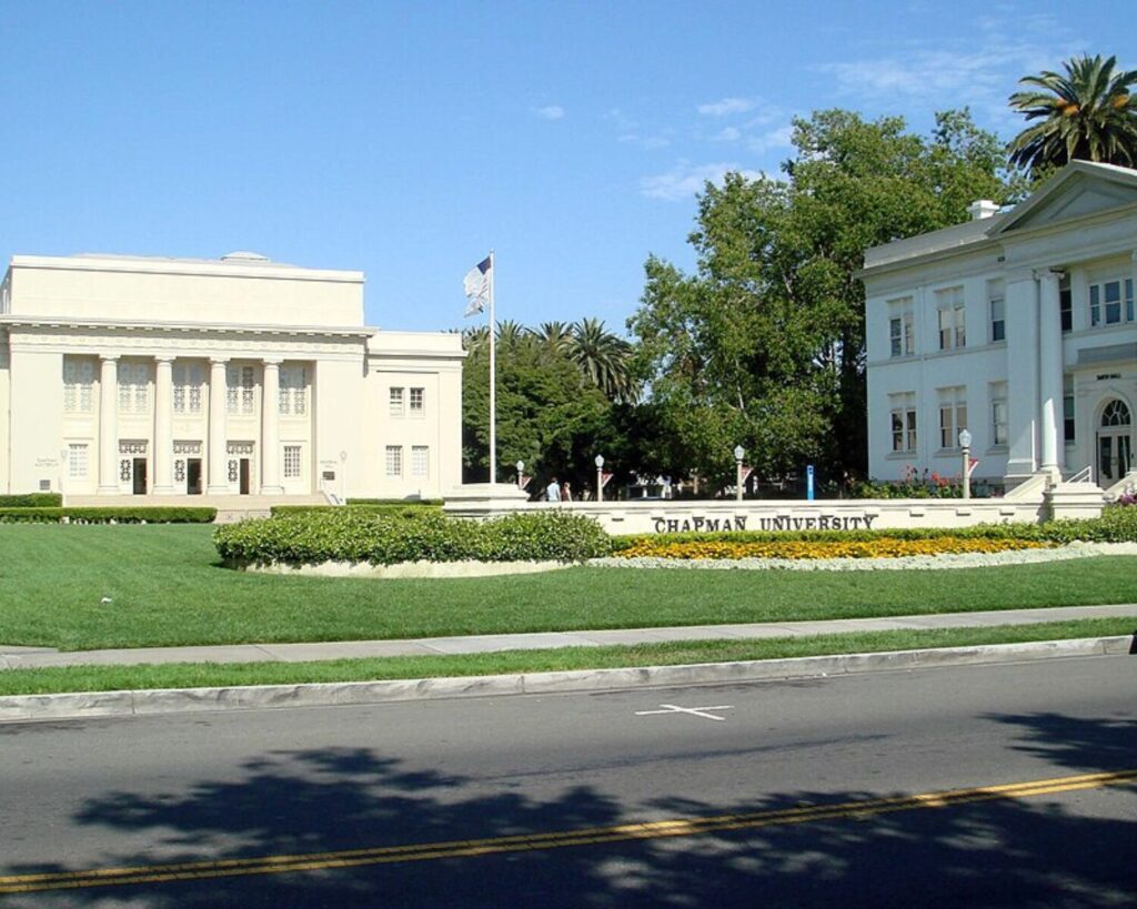 Front view of Chapman University campus in Orange, California, featuring neoclassical academic buildings, green lawn, palm trees, and a street in the foreground under a clear blue sky.