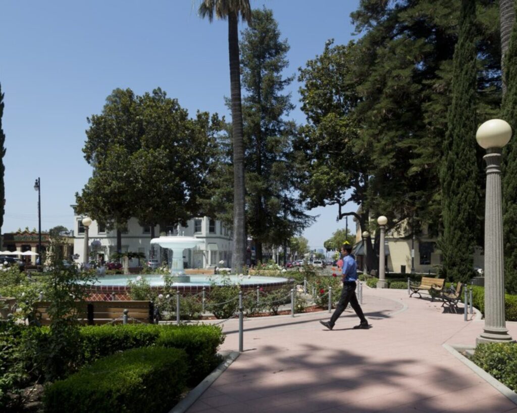 Park in the center of Old Town Orange, California, featuring a circular fountain, landscaped garden beds, tall trees, walking paths, and a person strolling through the plaza on a sunny day.