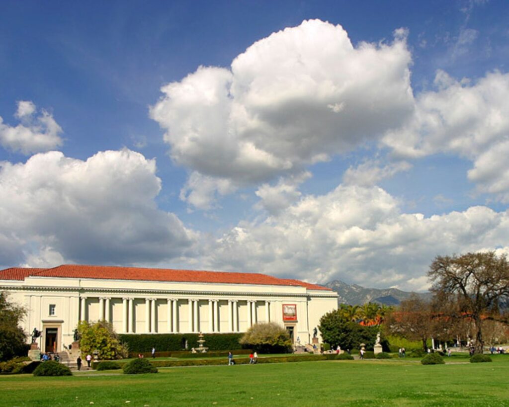 Huntington Library building with landscaped gardens, visitors, and mountains in background under cloudy sky in California