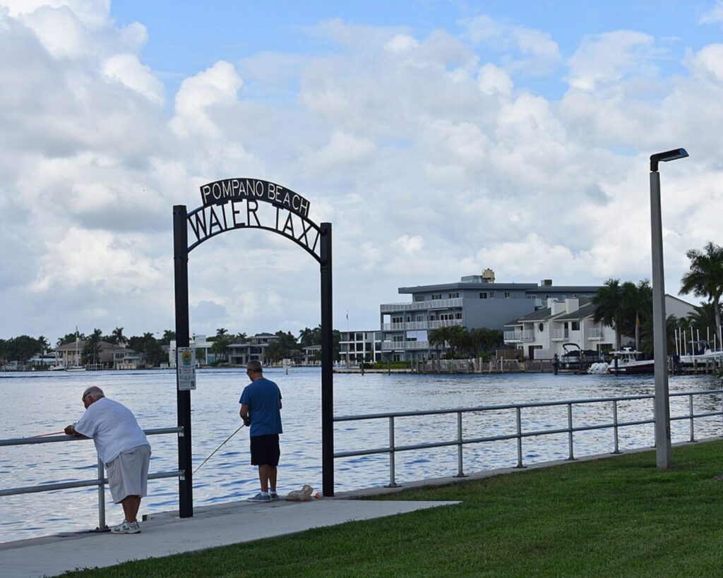 Entrance sign reading “Pompano Beach Waterway” at Indian Mound Park with two men fishing by the waterfront, surrounded by residential buildings and palm trees under a cloudy sky