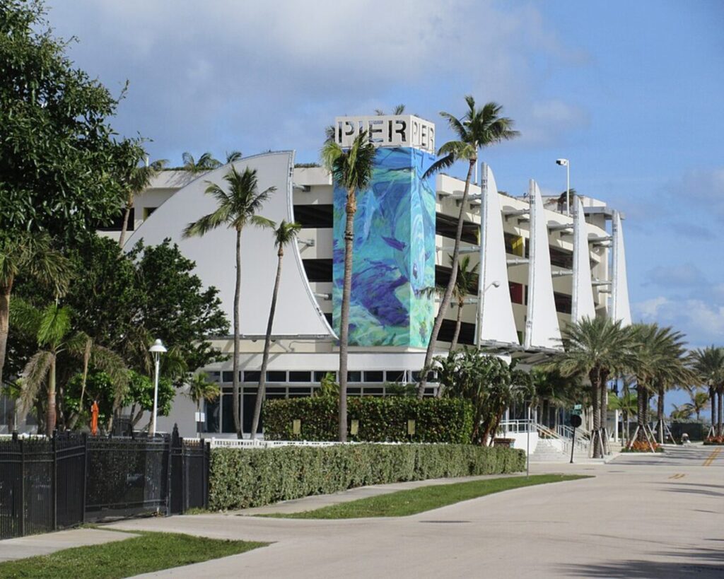 Modern Pompano Beach pier parking structure with a tall cylindrical tower featuring a colorful ocean-themed mural, surrounded by palm trees and a clear sky