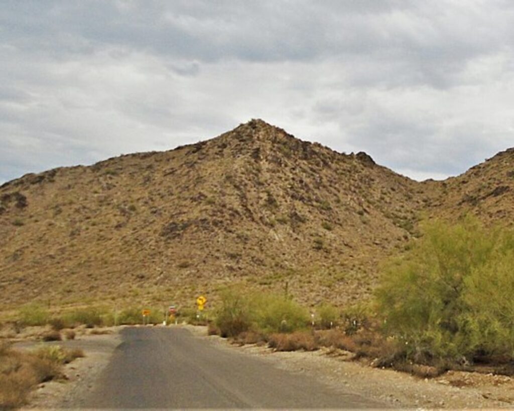 Desert landscape with a narrow paved road leading toward the San Tan Mountains, featuring rocky hills, sparse shrubs, and cloudy skies overhead.