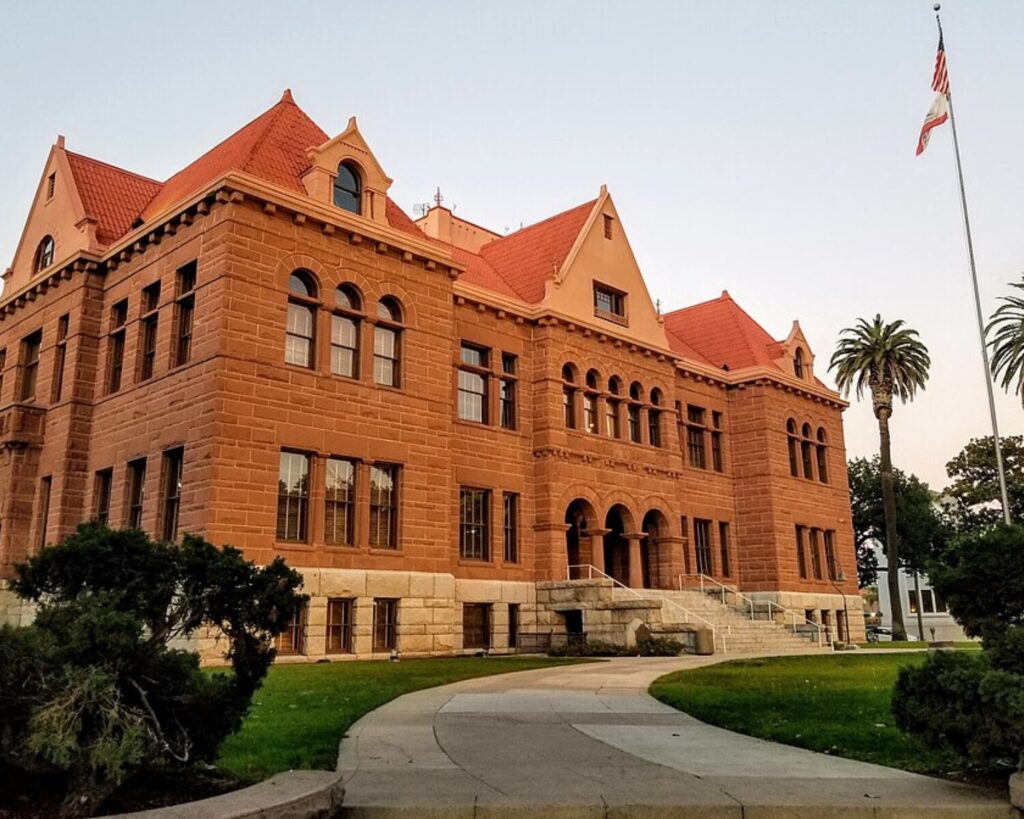 Historic Old Orange County Courthouse in Santa Ana, California, featuring a red-brick building with arched windows, a red-tiled roof, landscaped lawn, and a flagpole with the American flag nearby.
