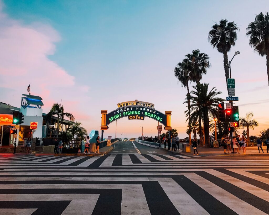 Wide zebra crossing leading to the Santa Monica Pier entrance sign at sunset, with palm trees, traffic lights, and pedestrians on both sides under a colorful evening sky.