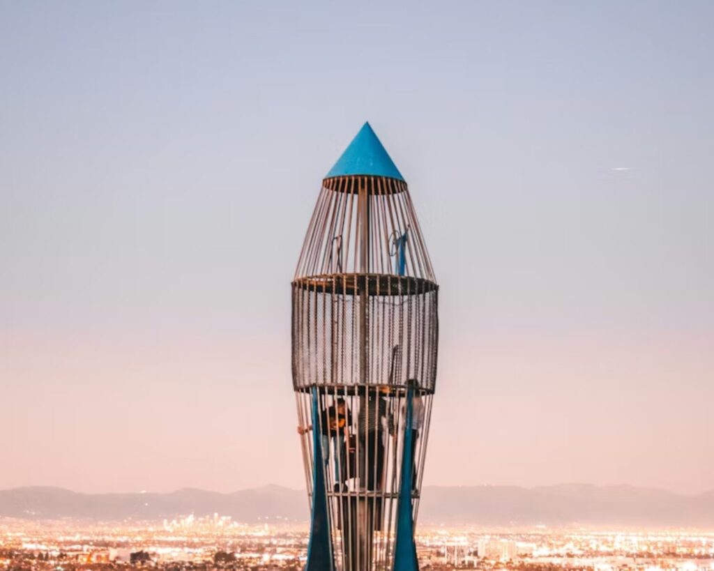 Rocket-shaped metal sculpture at Los Arboles Rocketship Park overlooking a cityscape at dusk with lights glowing in the distance