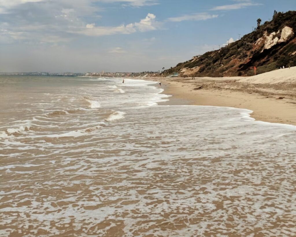 Waves washing onto a sandy beach with rocky cliffs along the shoreline under a partly cloudy sky during daytime