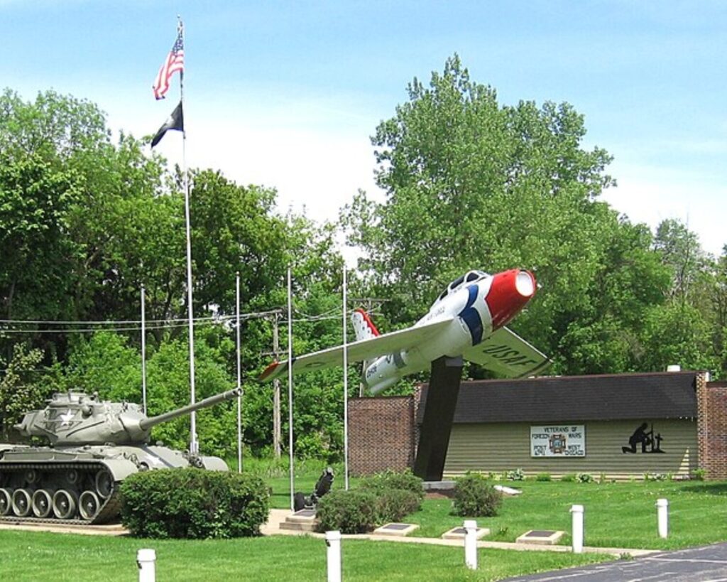 Military memorial display with a tank and mounted aircraft outside VFW Post building in West Chicago, Illinois, with American flag and green lawn