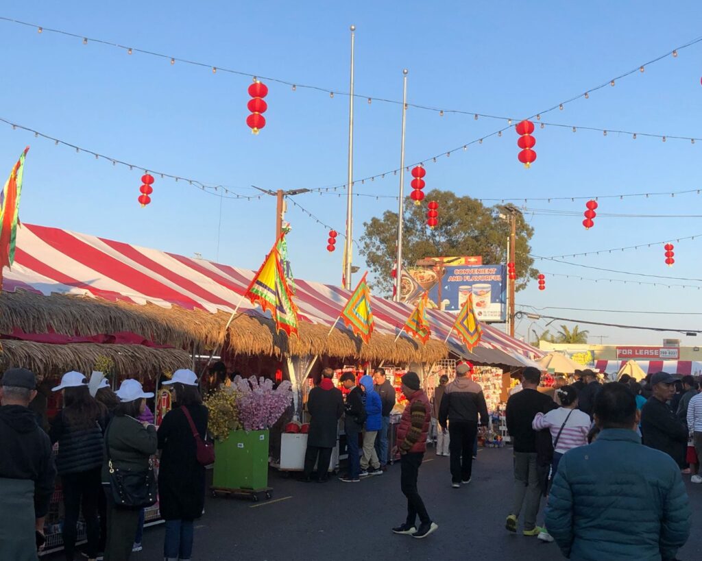 Crowd walking through outdoor street market with food stalls colorful flags and hanging red lanterns during festival
