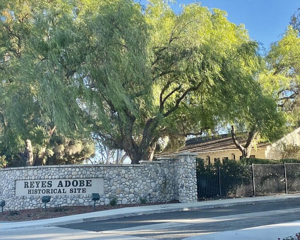 Stone entrance wall with a sign that reads 'Reyes Adobe Historical Site' and large green trees behind it.