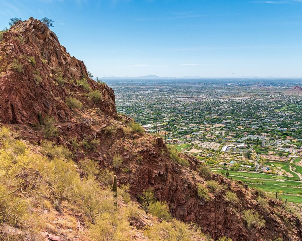 Panoramic view from Camelback Mountain in Phoenix, Arizona, featuring rugged red rock cliffs and desert vegetation in the foreground overlooking the sprawling cityscape below under a clear blue sky.
