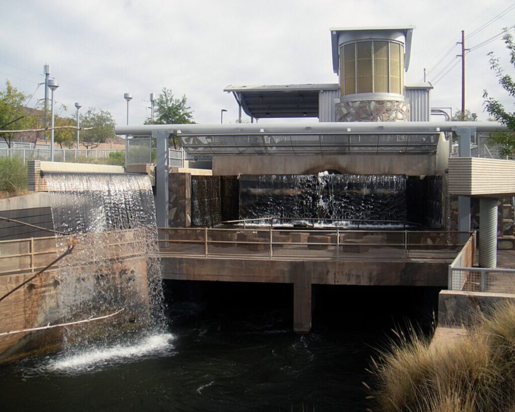 A modern urban waterfall feature at Arizona Falls in Arizona, showing cascading water flowing over concrete ledges into a dark pool below. The structure includes metal railings, stone accents, and a cylindrical observation tower under an overcast sky, with desert-style landscaping surrounding the area.