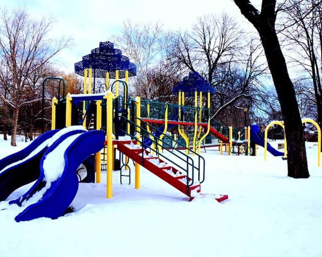 Colorful playground at Proksa Park covered in snow, featuring slides, climbing structures, and bare trees in winter.