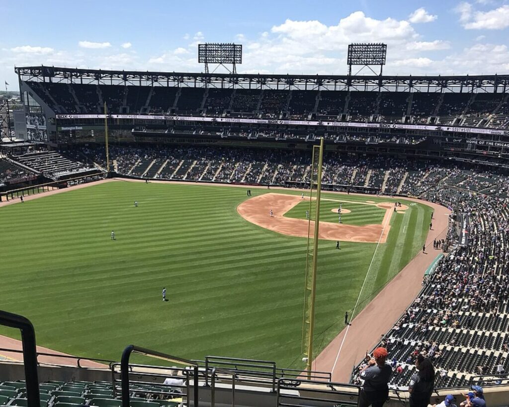 Wide view inside Guaranteed Rate Field during a Chicago White Sox and New York Mets baseball game, showing the green baseball diamond, players on the field, packed stadium seating, and large scoreboard displays under a bright sky.