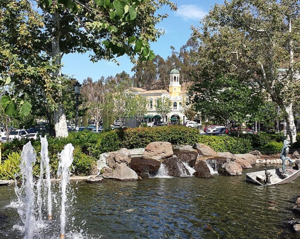 Landscaped pond with fountain jets, rocks, and trees at Calabasas Commons shopping area in Calabasas, California, with storefronts and parked cars in the background.