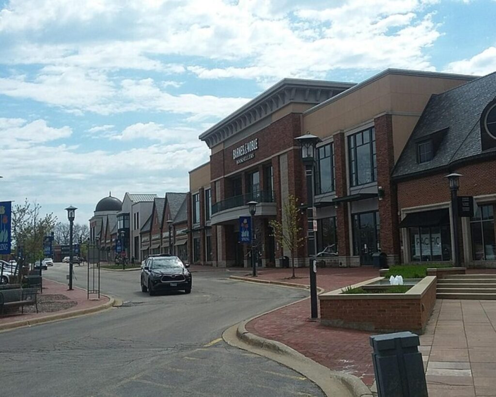 Deerfield Square shopping district with brick storefronts, parked cars, and a curved street lined with lampposts under a partly cloudy sky.