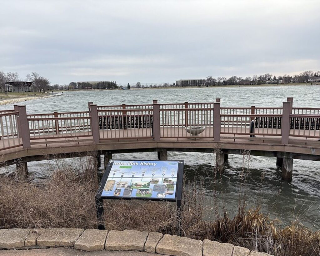 Boardwalk overlook at Lake Opeka in Des Plaines, Illinois, with railings, lake views, and an informational history sign under cloudy skies.
