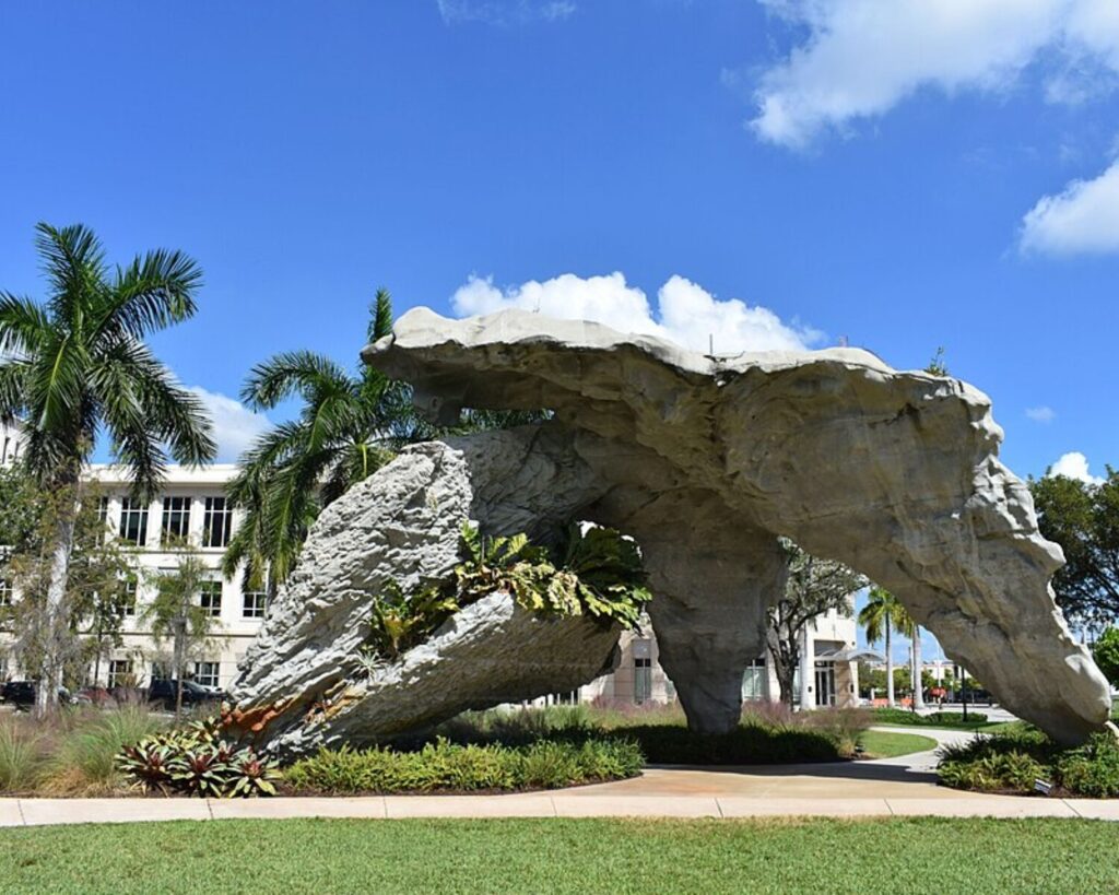 Large stone arch sculpture in a landscaped campus courtyard with palm trees and a white building in the background under a blue sky.