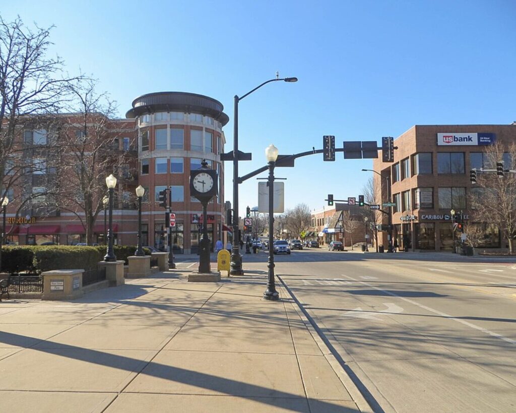 City street intersection with brick buildings, a prominent clock on a stand, street lamps, and light traffic on a sunny day.
