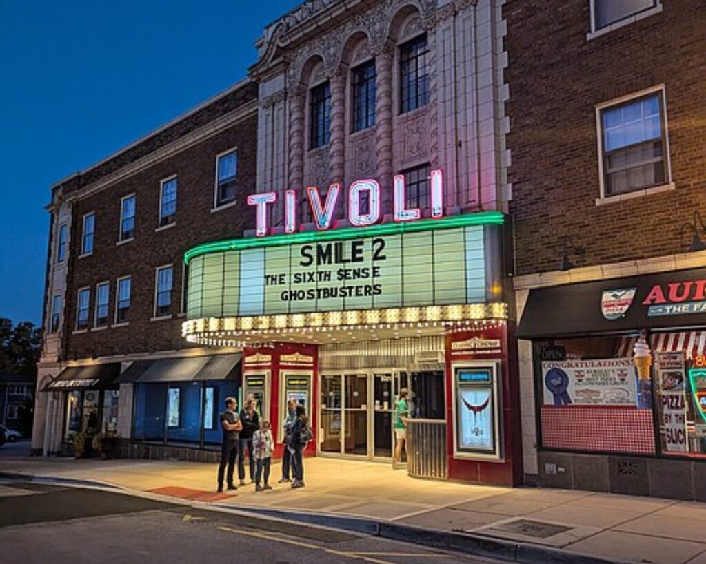 Historic Tivoli Theatre in downtown Downers Grove at dusk, featuring a glowing neon marquee, brick facade, and pedestrians standing outside the entrance.