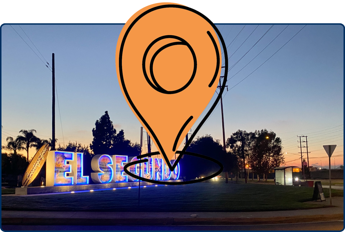 Illuminated “El Segundo” welcome sign at dusk, surrounded by palm trees, power lines, and a roadway under a fading evening sky.