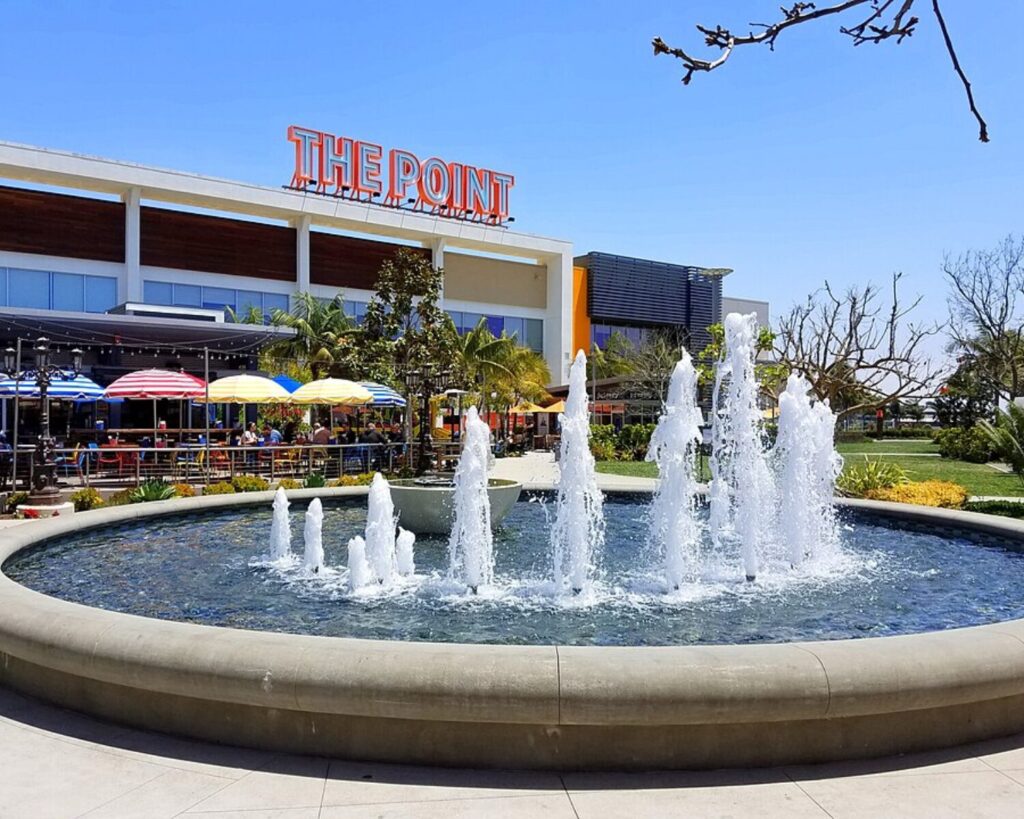 Outdoor shopping and dining area at The Point in El Segundo, California, featuring a large circular fountain, colorful umbrellas, and modern storefronts under a clear blue sky.