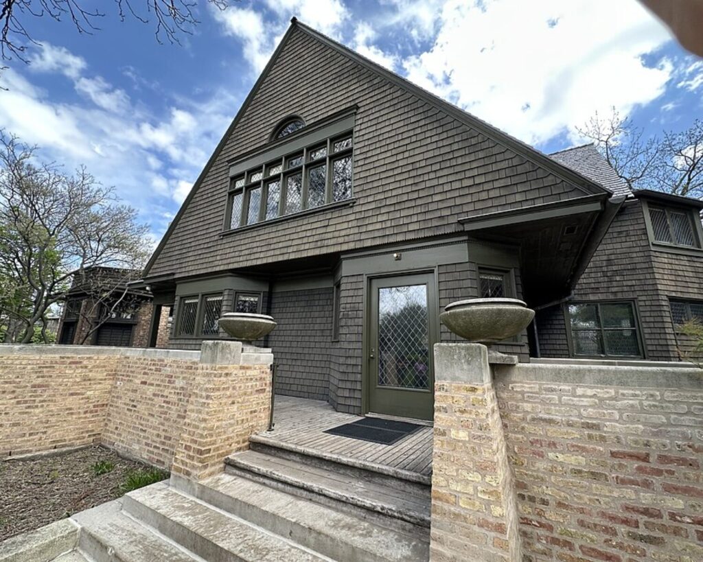 Front entrance of a dark-gray shingle house with a brick boundary wall, concrete steps, and two stone planters flanking a lattice-door entry.