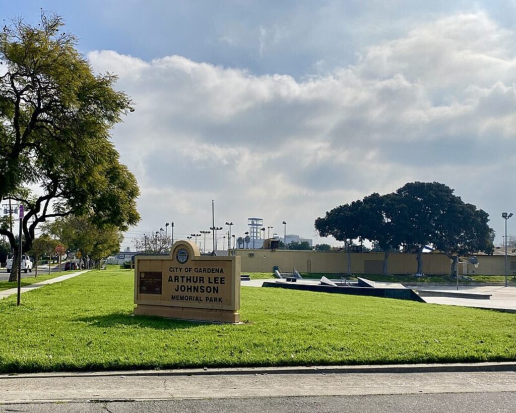 Grassy park area in Gardena, California, with a memorial park sign, trees, sidewalks, and recreational facilities under a partly cloudy sky.