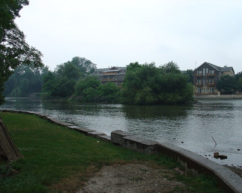 Calm stretch of the Fox River in Geneva, Illinois, bordered by trees, grassy riverbank paths, and residential buildings along the opposite shore.