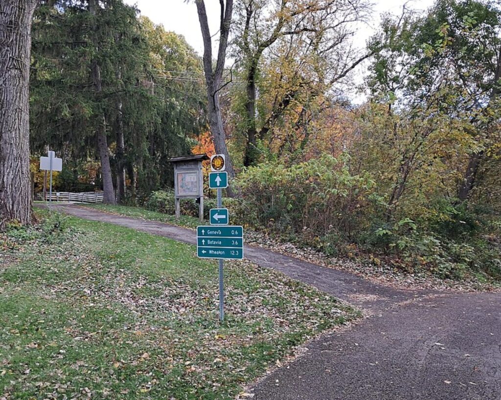Forked section of the Fox River Trail in Geneva, Illinois, surrounded by autumn trees and greenery, with directional trail signs along the paved path.