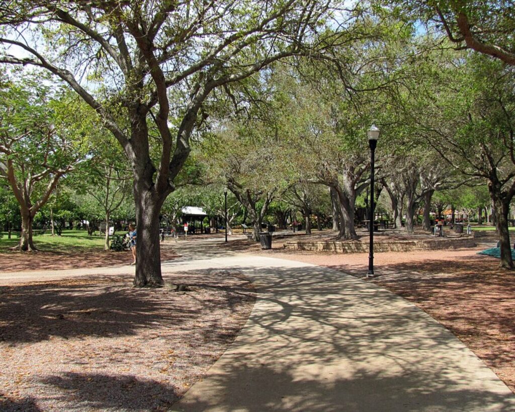 Curved walking path through Largo Central Park in Florida, surrounded by large shaded trees, benches, and lamp posts on a sunny day.