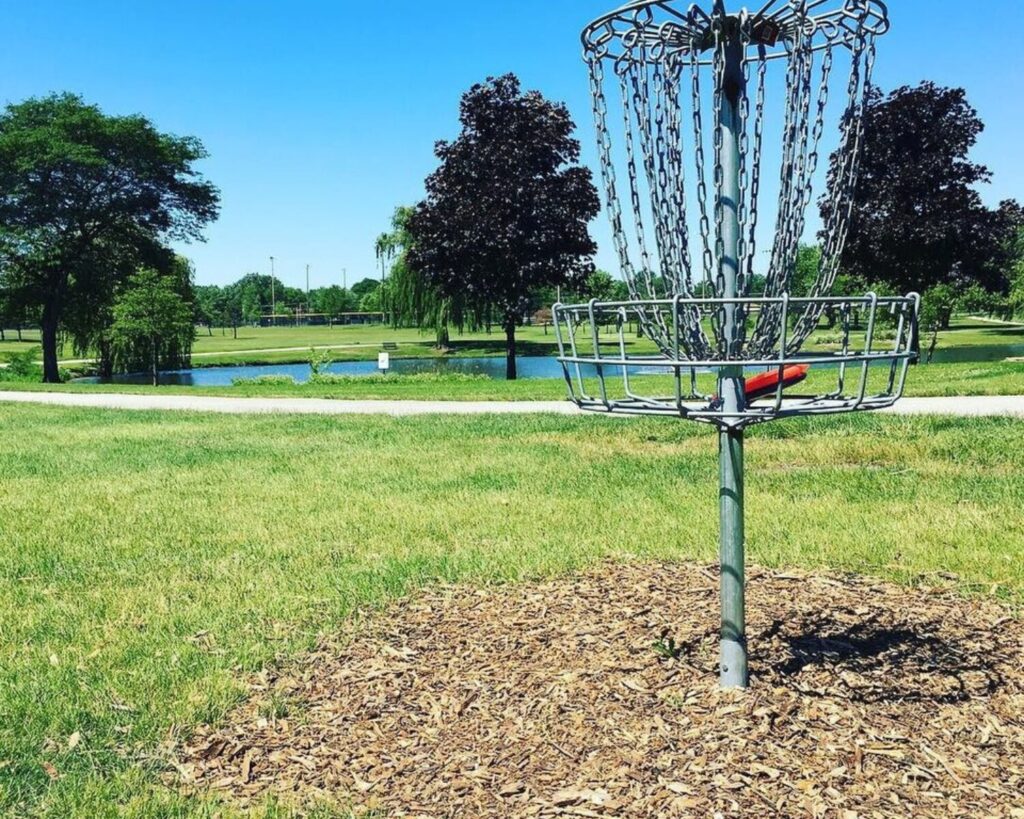 Disc golf basket at Madison Meadow Disc Golf Course in Lombard, Illinois, surrounded by grassy parkland, trees, and a small pond.