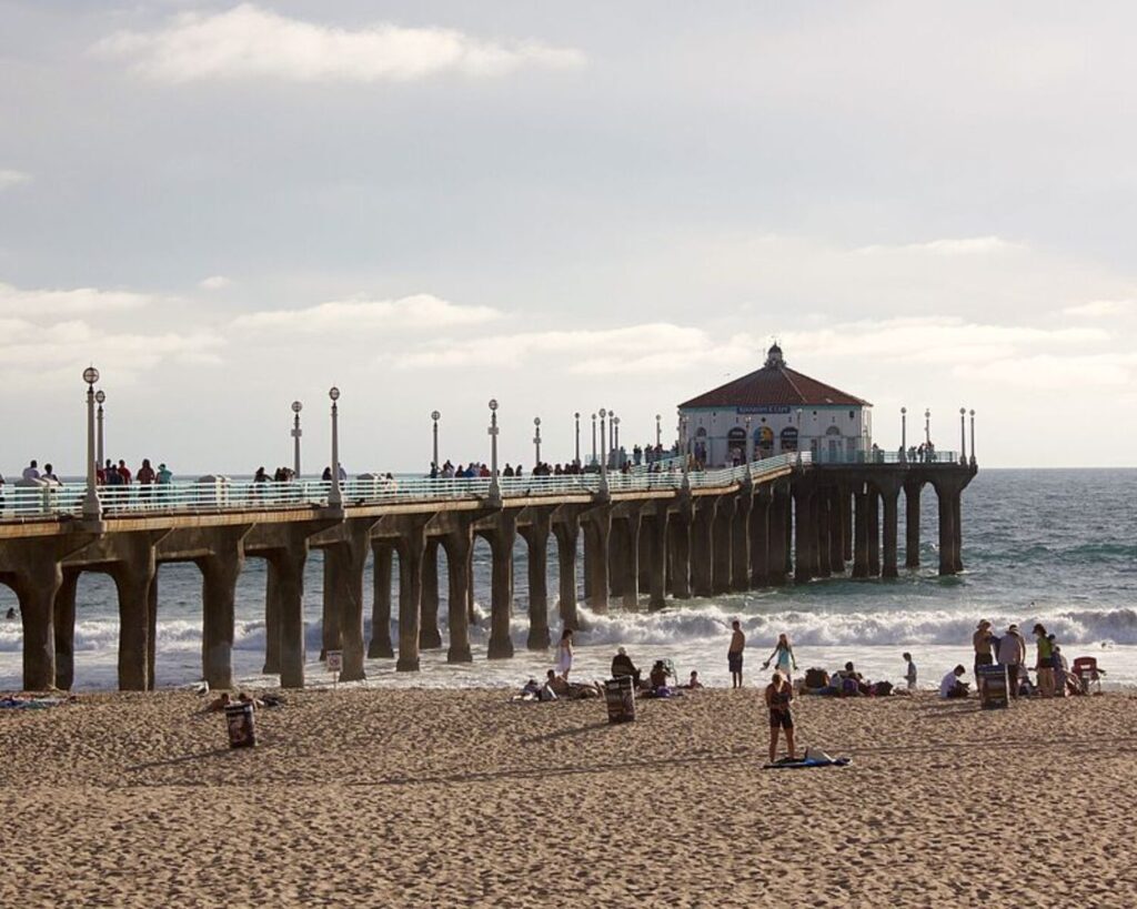 Manhattan Beach Pier extending into the Pacific Ocean with beachgoers relaxing on the sandy shore and people walking along the pier under a hazy coastal sky.