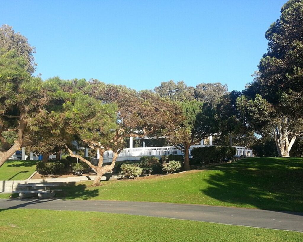 Sunlit park with green lawn, several trees, and a white railing along a path in the background.