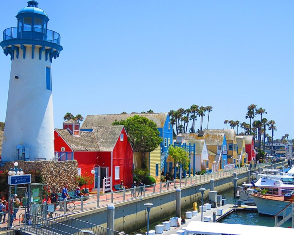 White lighthouse beside colorful waterfront buildings and docked boats along the marina in Marina del Rey, Los Angeles, California, under a clear blue sky.